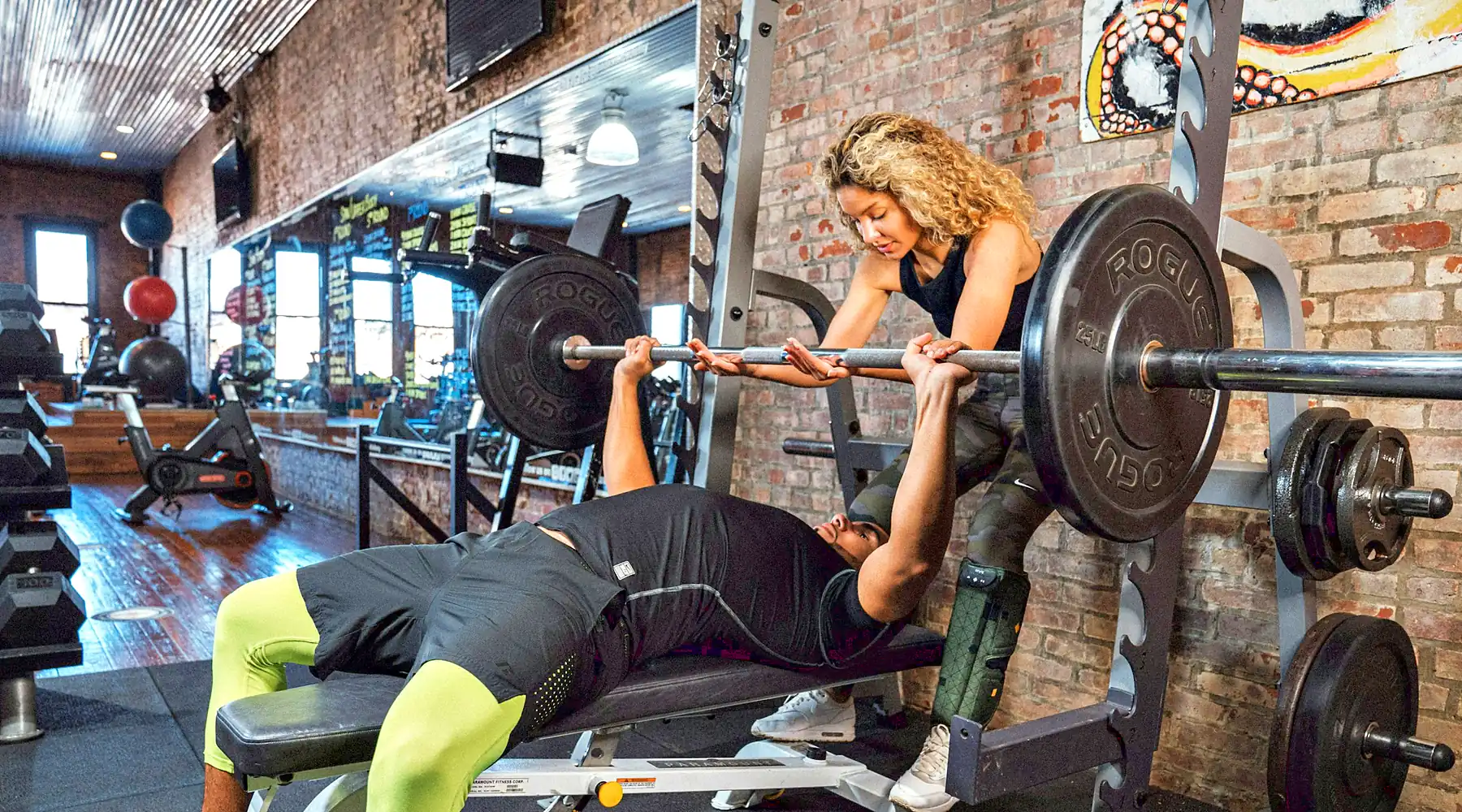 A woman spots a lifter on the bench press in a brick-walled gym